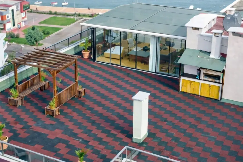 Kırmızı ve koyu renkli karo kauçukla kaplı çatı terası: cam bölmeli bir köşk, ahşap pergola, saksılar ve arkada deniz